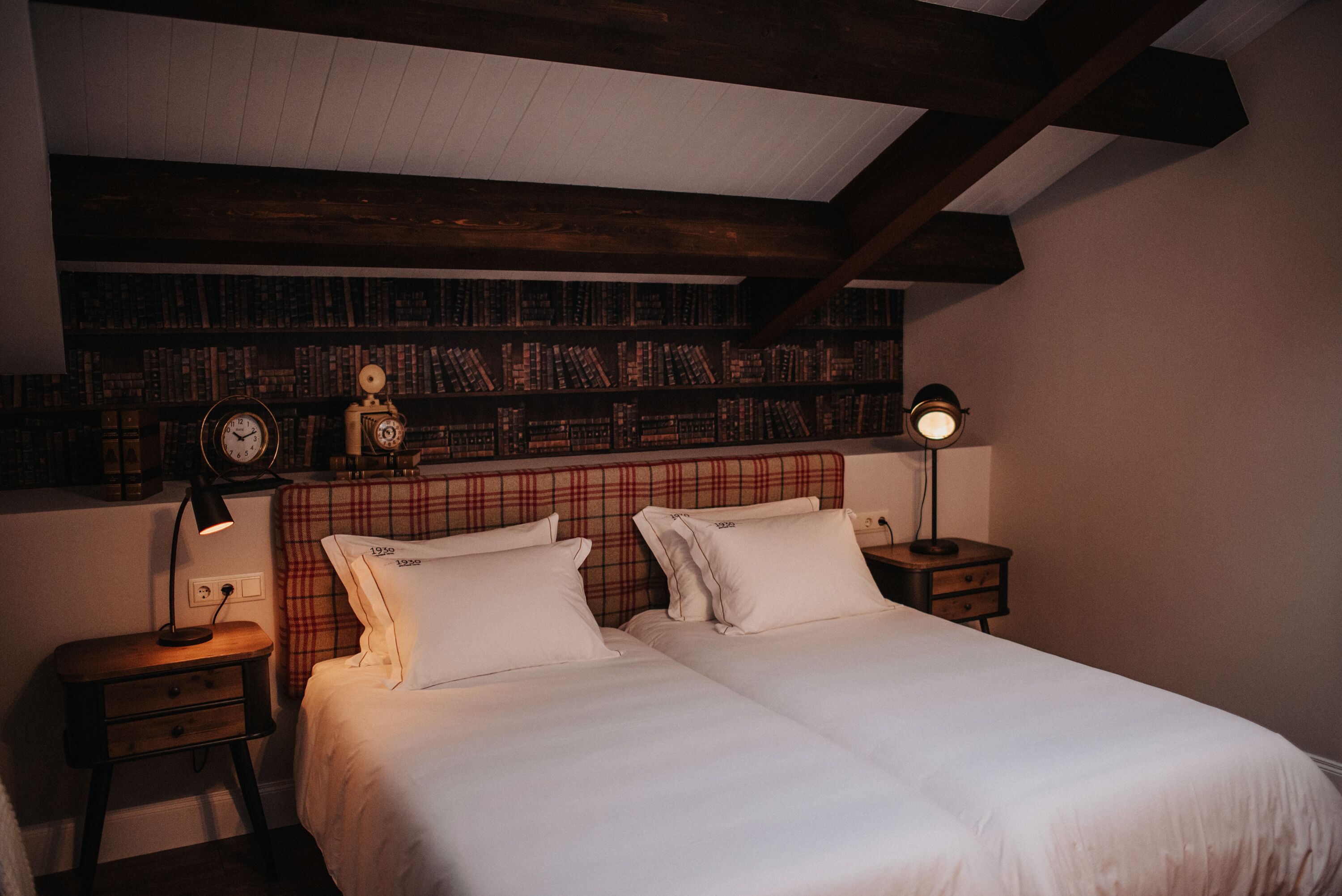 A bedroom with two beds, a bookshelf, a clock, and a nightstand with a lamp.