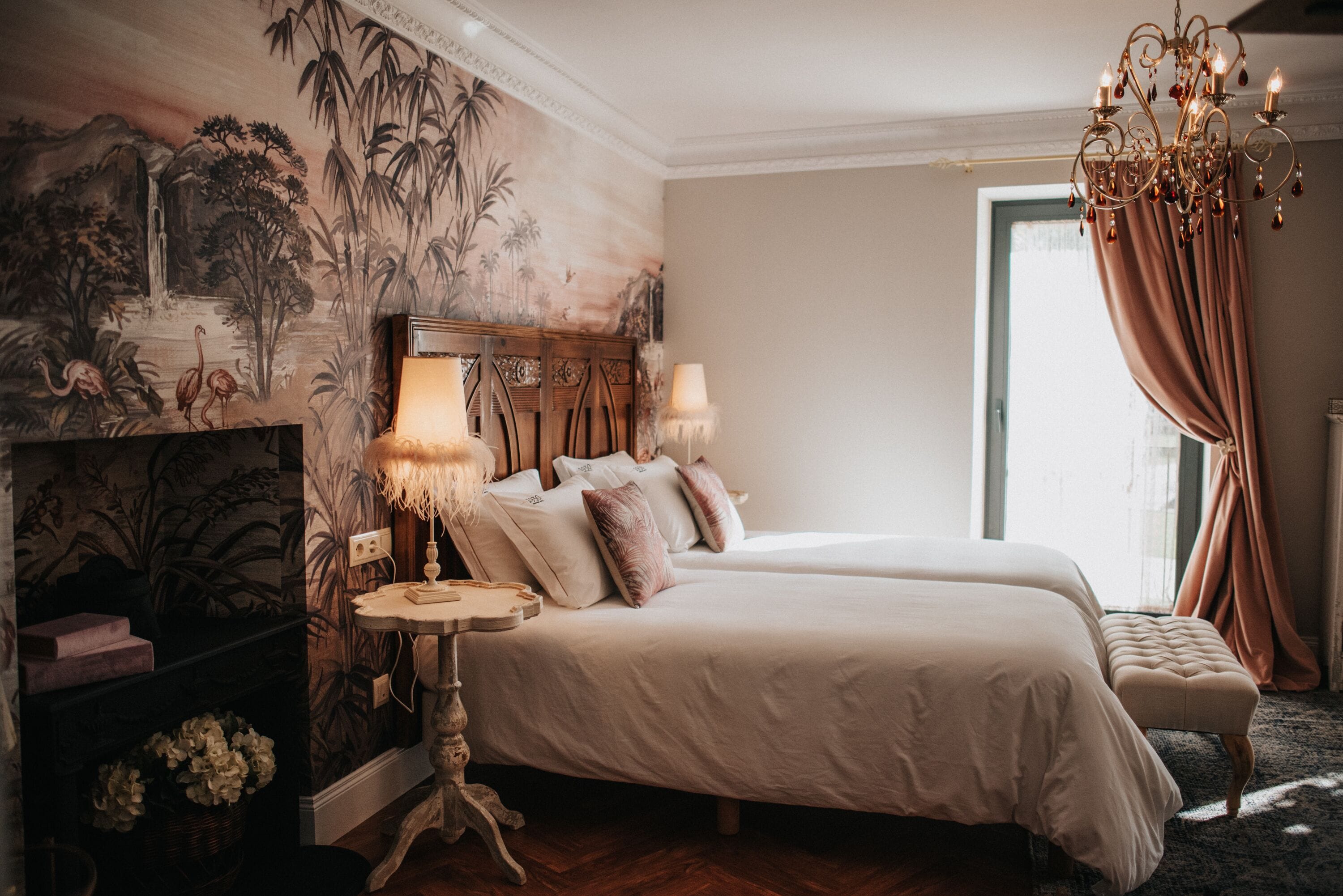Une chambre avec deux lits, un lustre, une fresque murale et une fenêtre avec des rideaux.