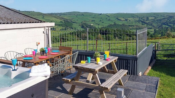 A patio with a wooden table and chairs, a hot tub, and a view of rolling hills. - 4 bedroom accommodation in Nantglyn, near Denbigh (Nantglyn, near Denbigh)