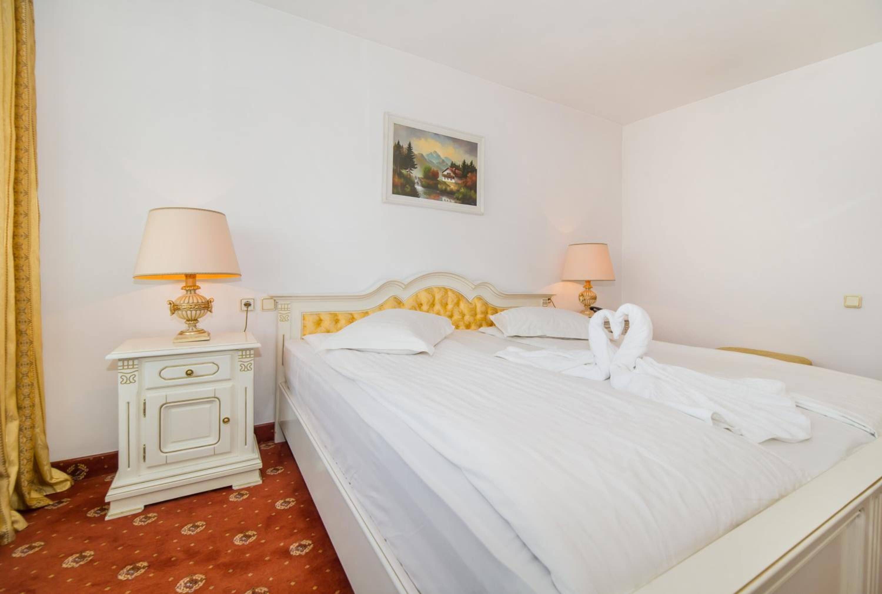A neatly made bed with white linens and two towels, a bedside table with a lamp, and a painting on the wall.