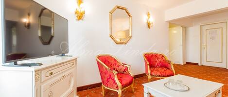 A hotel room with a TV, two red upholstered chairs, a white table, and a patterned carpet.