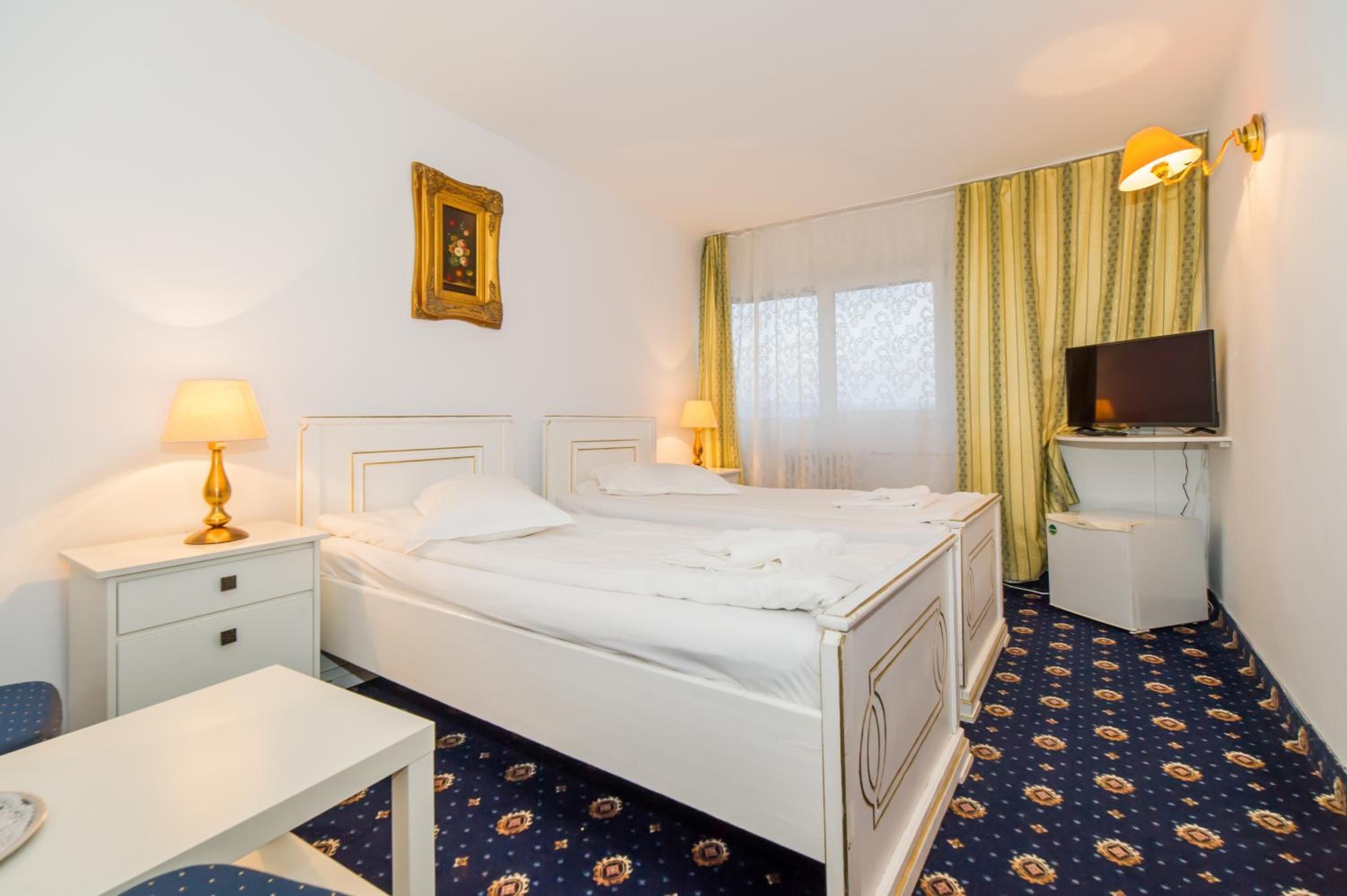 A hotel room with a bed, bedside tables, a desk, a television, and a window with curtains.