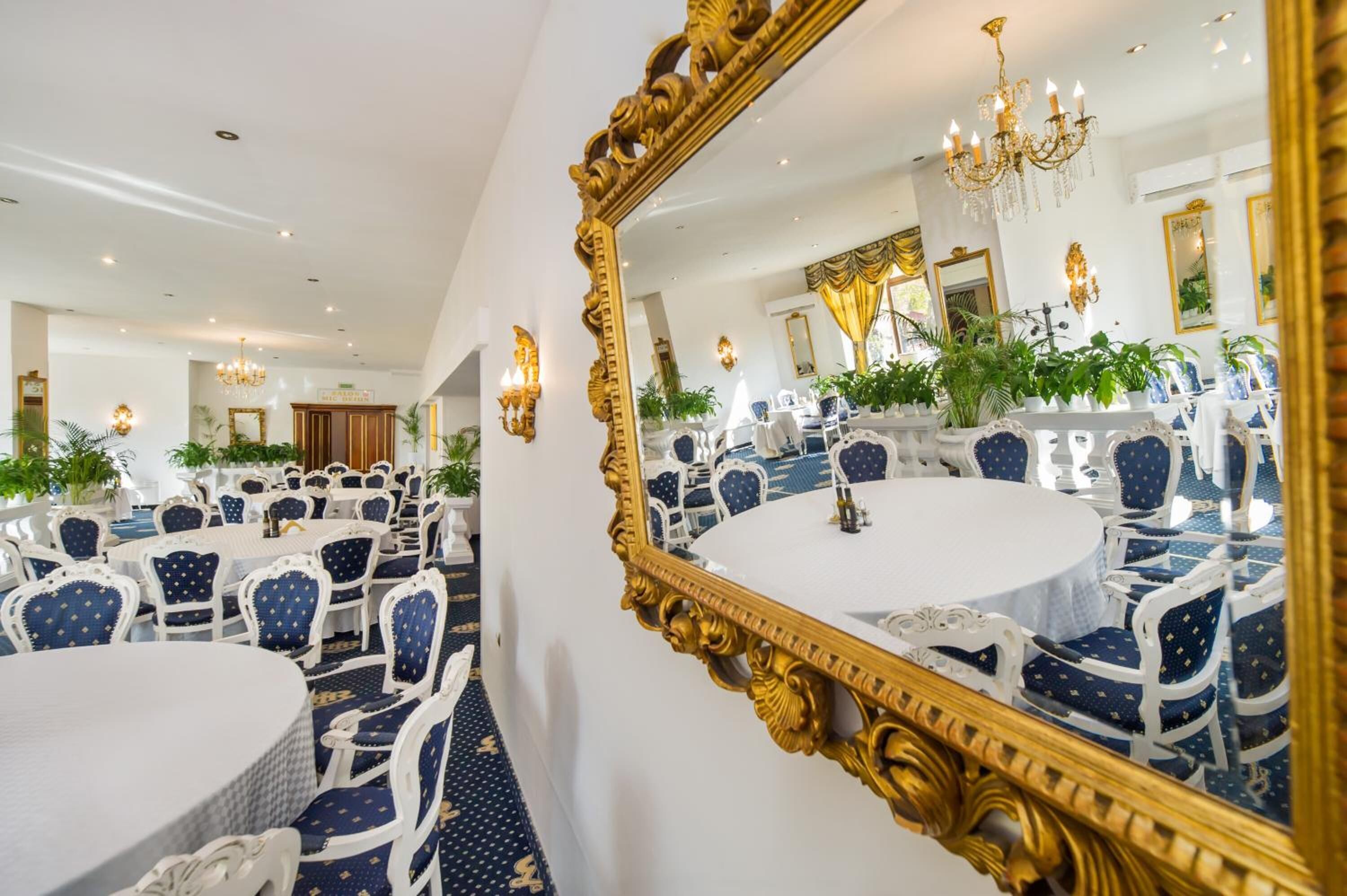 A formal dining room with round tables and blue upholstered chairs, a large ornate mirror, and a chandelier.