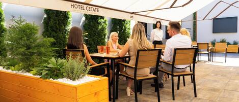 A covered outdoor seating area with wooden tables and chairs, surrounded by greenery and a sign reading "HOTEL CETATE."