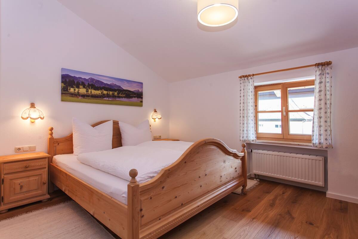 Une chambre à coucher avec un lit en bois, deux tables de chevet, une commode, une fenêtre avec des rideaux, un radiateur et un tableau accroché au mur.