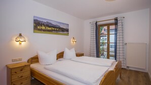 A bedroom with a wooden bed, two bedside tables, a nightstand, a wall-mounted lamp, a window with a view of a mountain landscape, and a radiator. - Schönes Ammertal (Oberammergau)