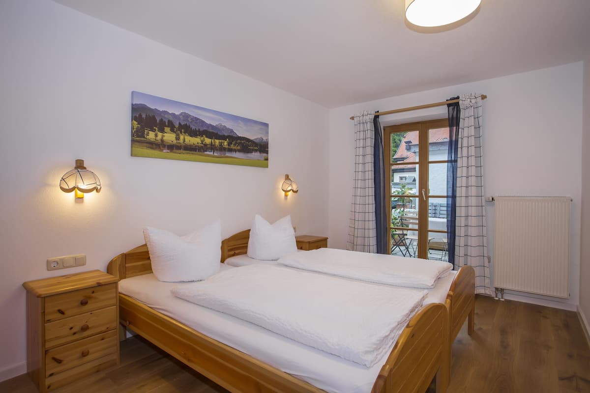 Une chambre à coucher avec un lit en bois, deux tables de chevet, une commode, une lampe fixée au mur, une fenêtre donnant sur un paysage montagneux et un radiateur.