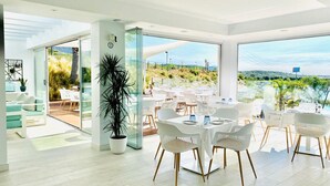 A modern dining area with white tables and chairs, large windows offering a view of the landscape, and a potted plant in the center. - Exclusive 3-bed Penthouse Alcazaba Lagoon (321) (Casares)