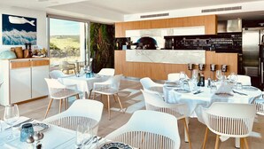 A modern dining area with white chairs and tables, a kitchen in the background, and a large window with a view of the outdoors. - Exclusive 3-bed Penthouse Alcazaba Lagoon (321) (Casares)