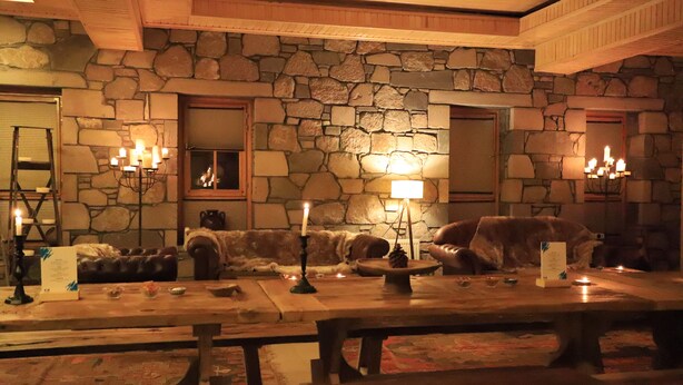 Una habitación acogedora con paredes de piedra, techo de madera y chimenea de piedra. Hay velas sobre la mesa y un plaid de piel en el sofá.