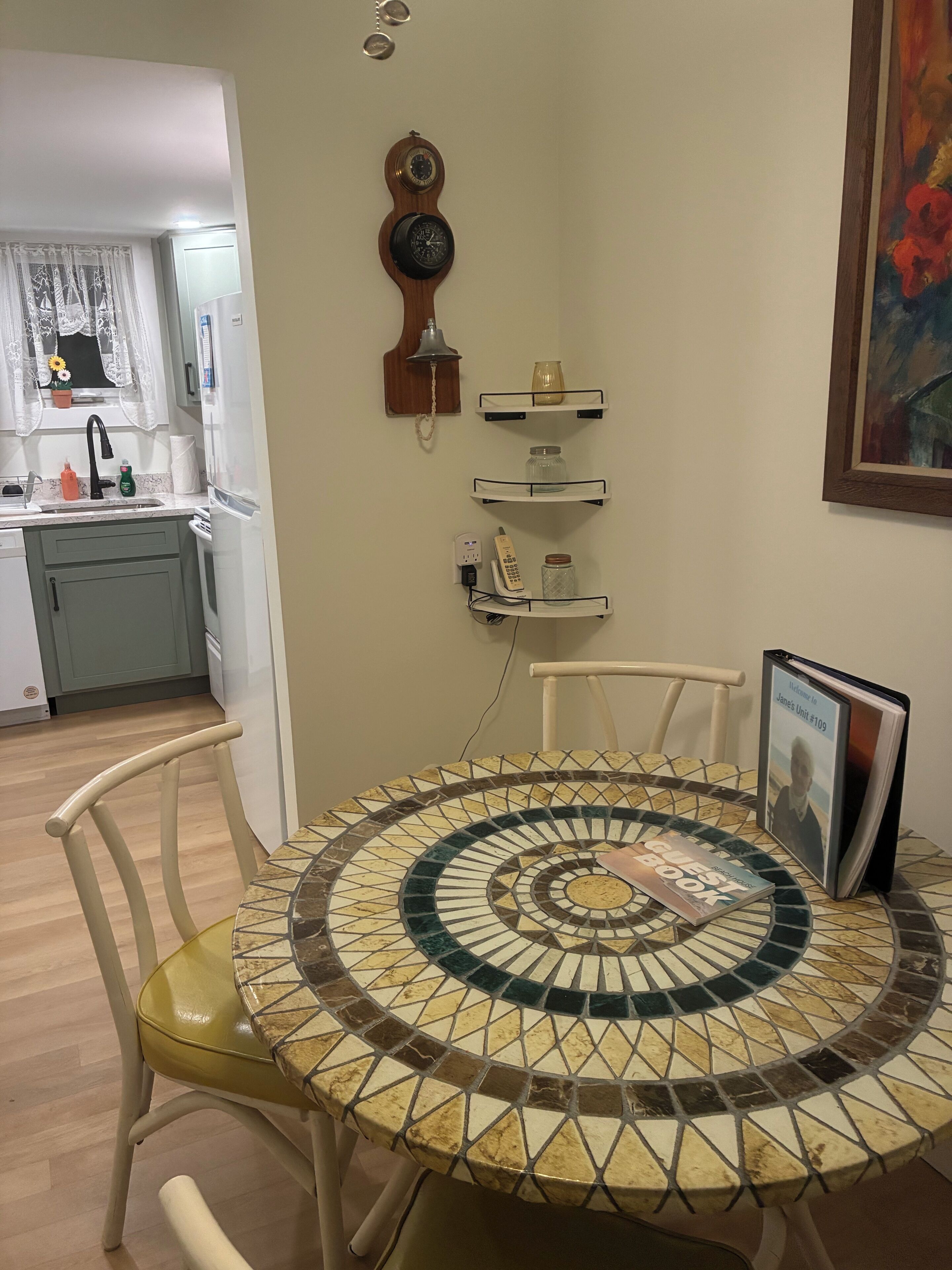 A dining area with a mosaic table, chairs, a framed welcome sign, and a kitchen in the background.