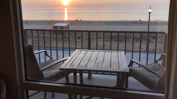 A balcony with a wooden table and two chairs, overlooking a beach with a sunset. - Flood Damaged - Renovated 2026 ~ Excuse Glitches - Still Quaint VINTAGE Studio (Rehoboth Beach)