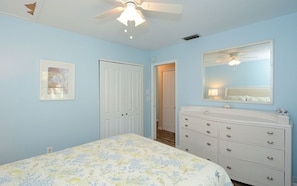 A bedroom with a bed, dresser, ceiling fan, and a door leading to another room.