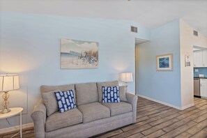 A living room with a sofa, two lamps, a painting, and a kitchen area.