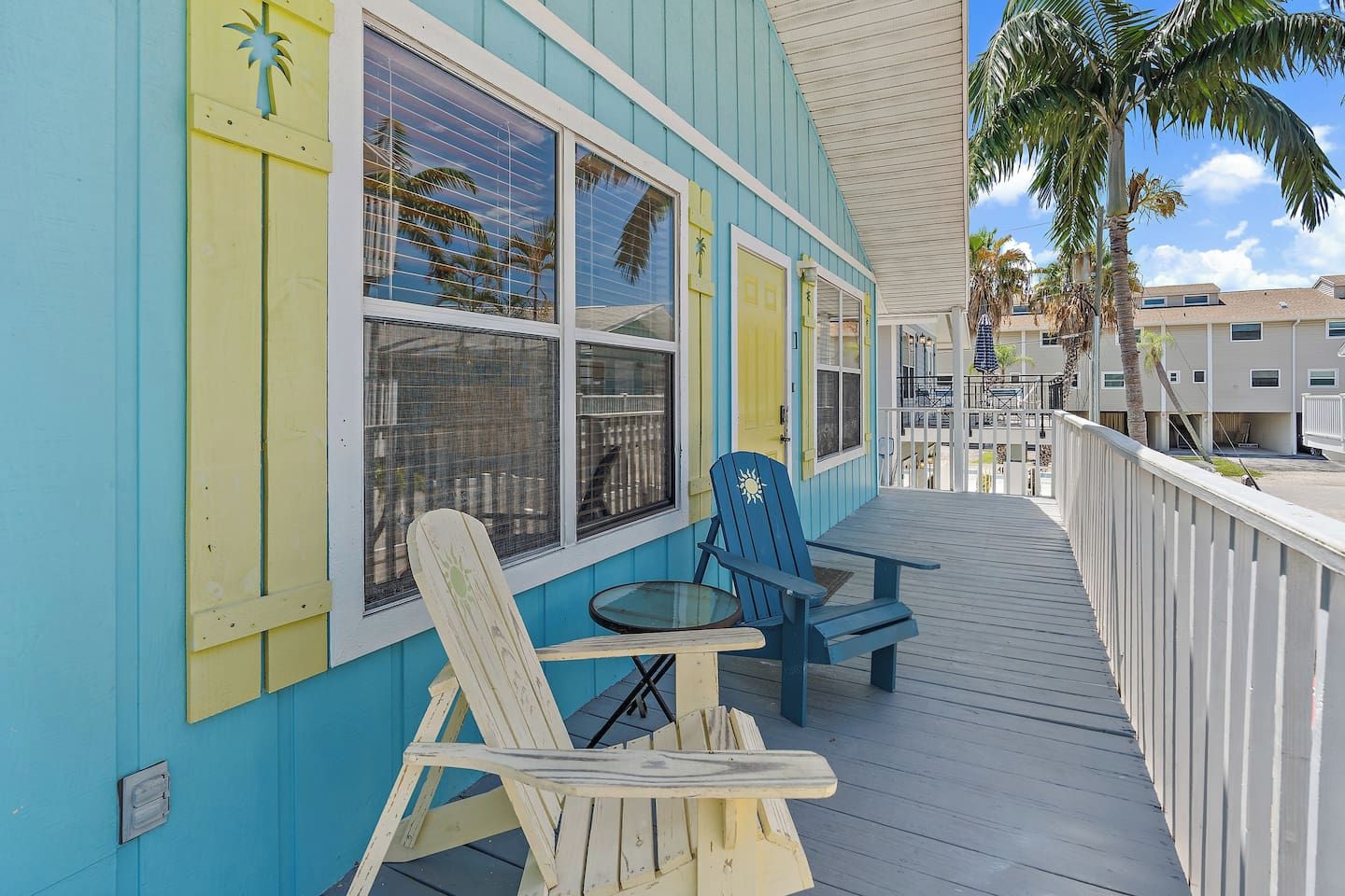 A wooden deck with two Adirondack chairs and a small table, overlooking a residential area with palm trees and a clear sky.