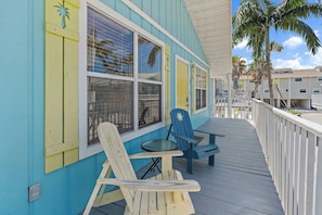 A wooden deck with two Adirondack chairs and a small table, overlooking a residential area with palm trees and a clear sky.