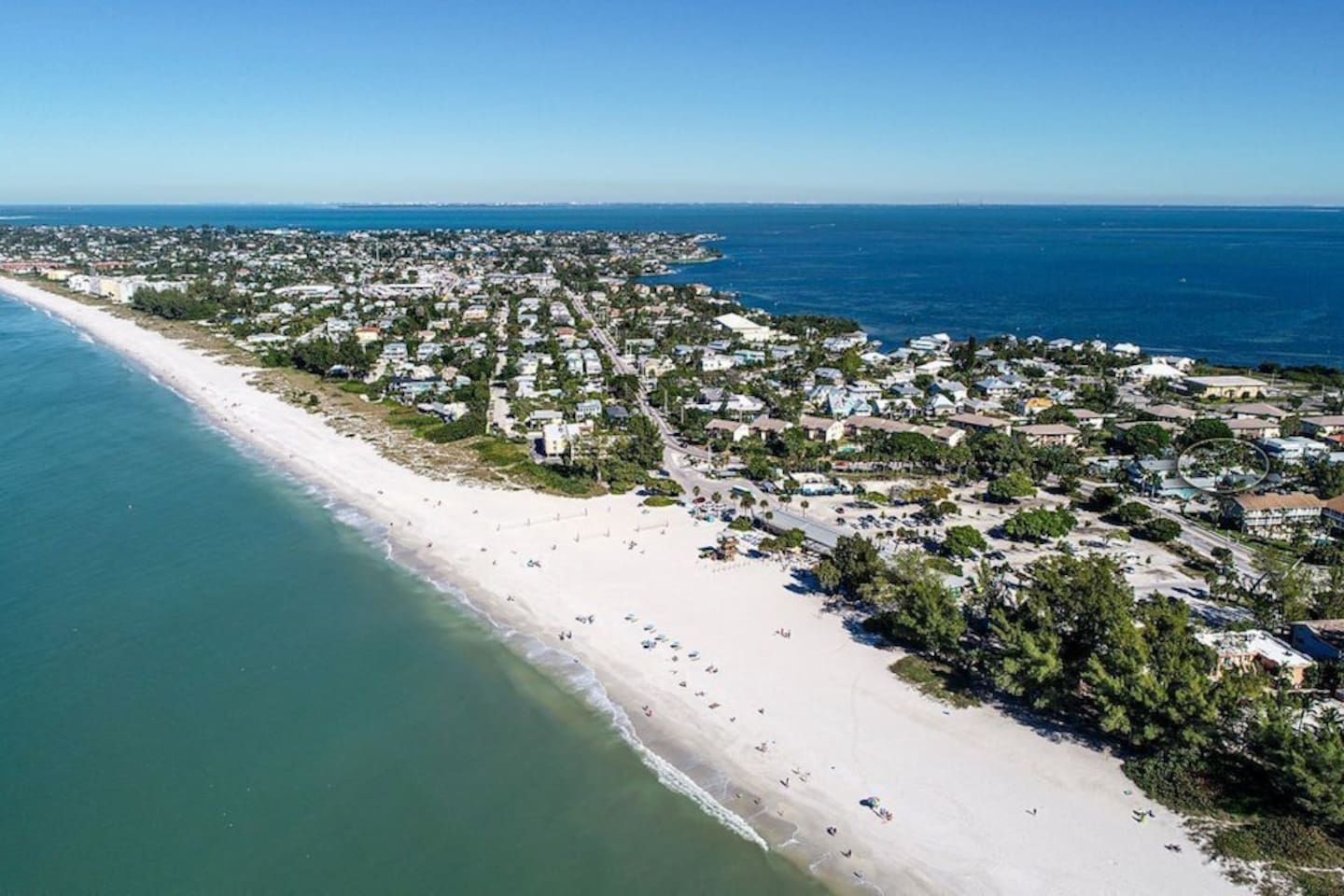 An aerial view of a coastal town with a long sandy beach, dense residential area, and clear blue sea.