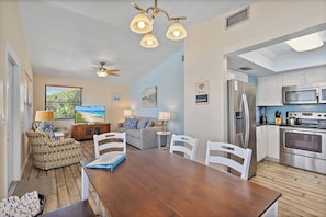 A modern kitchen with stainless steel appliances, a dining area with a wooden table and chairs, and a living room with a sofa and a television.