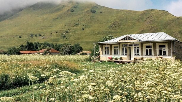 Una casa de exterior blanco con techo inclinado, rodeada por un campo de flores blancas, con una montaña al fondo.