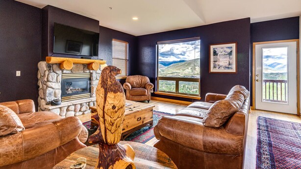 A living room with leather sofas, a stone fireplace, a large window with a mountain view, and a mounted television. - Exceptional Views, Large Townhome, Hot Tub, Pool, Private Garage 97A (Silverthorne)