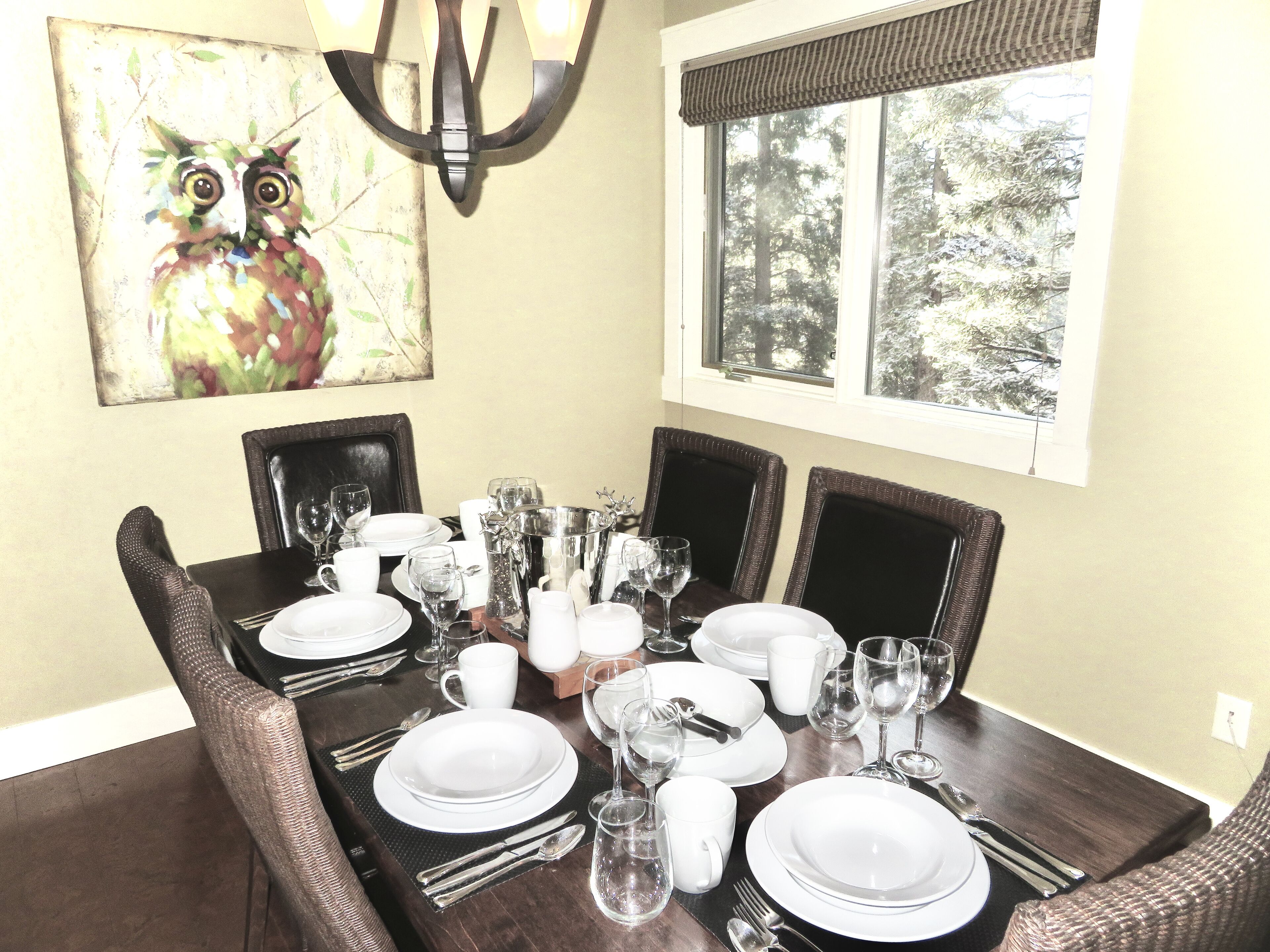 Une table à manger dressée avec de l’argenterie blanche, des verres et des couverts, entourée de chaises en osier, et ornée d’un tableau représentant une chouette au mur, ainsi que d’une fenêtre donnant sur un paysage arboré.