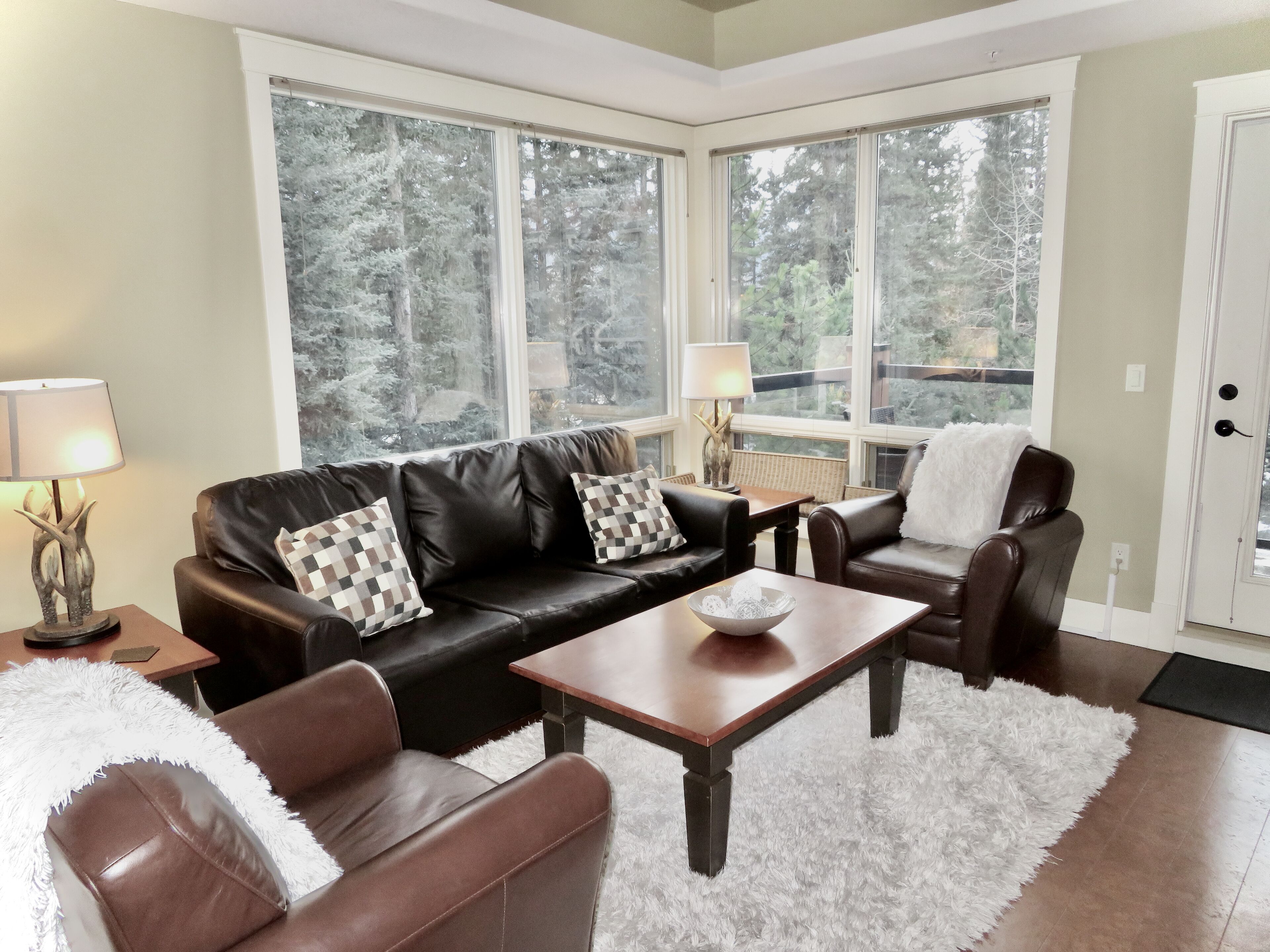 A living room with a brown leather sofa, a coffee table, and a white fluffy rug. There are two windows with a view of trees.