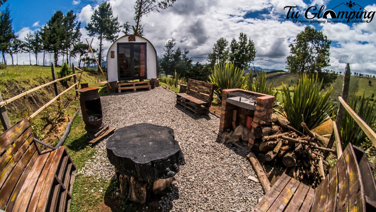 Un área de estar al aire libre de estilo rústico con bancos de madera y un brasero central, rodeada por un camino de grava y una valla de madera.