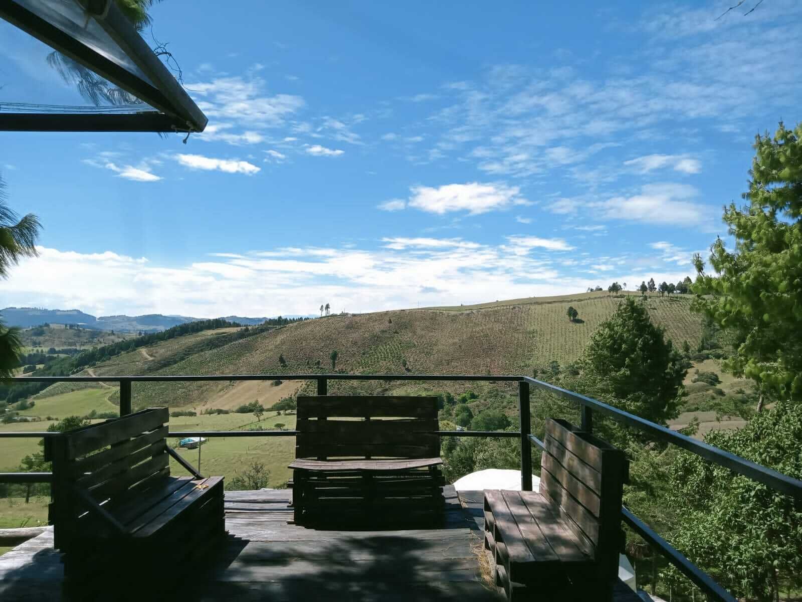 Ein Holzbänkchen und ein Tisch auf einem Balkon mit Blick auf eine hügelige Landschaft und einen klaren Himmel.