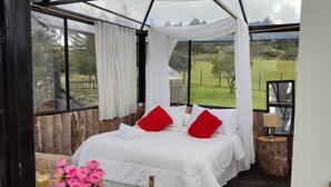 A bedroom with a large bed, white bedding, red pillows, a nightstand, a lamp, and a view of a green landscape through large windows. - Tu Glamping (Chocontá)