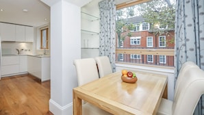 Een moderne keuken met witte kasten, een houten eettafel met een schaal fruit, en uitzicht op een rode baksteenbouw door een raam.