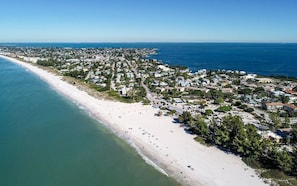 Luchtbeeld van een kustplaatsje met een lang zandstrand, een dichtbevolkt woongebied en helderblauw water.