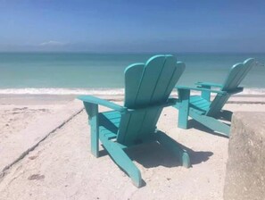 Two turquoise Adirondack chairs on a sandy beach with a view of the ocean.