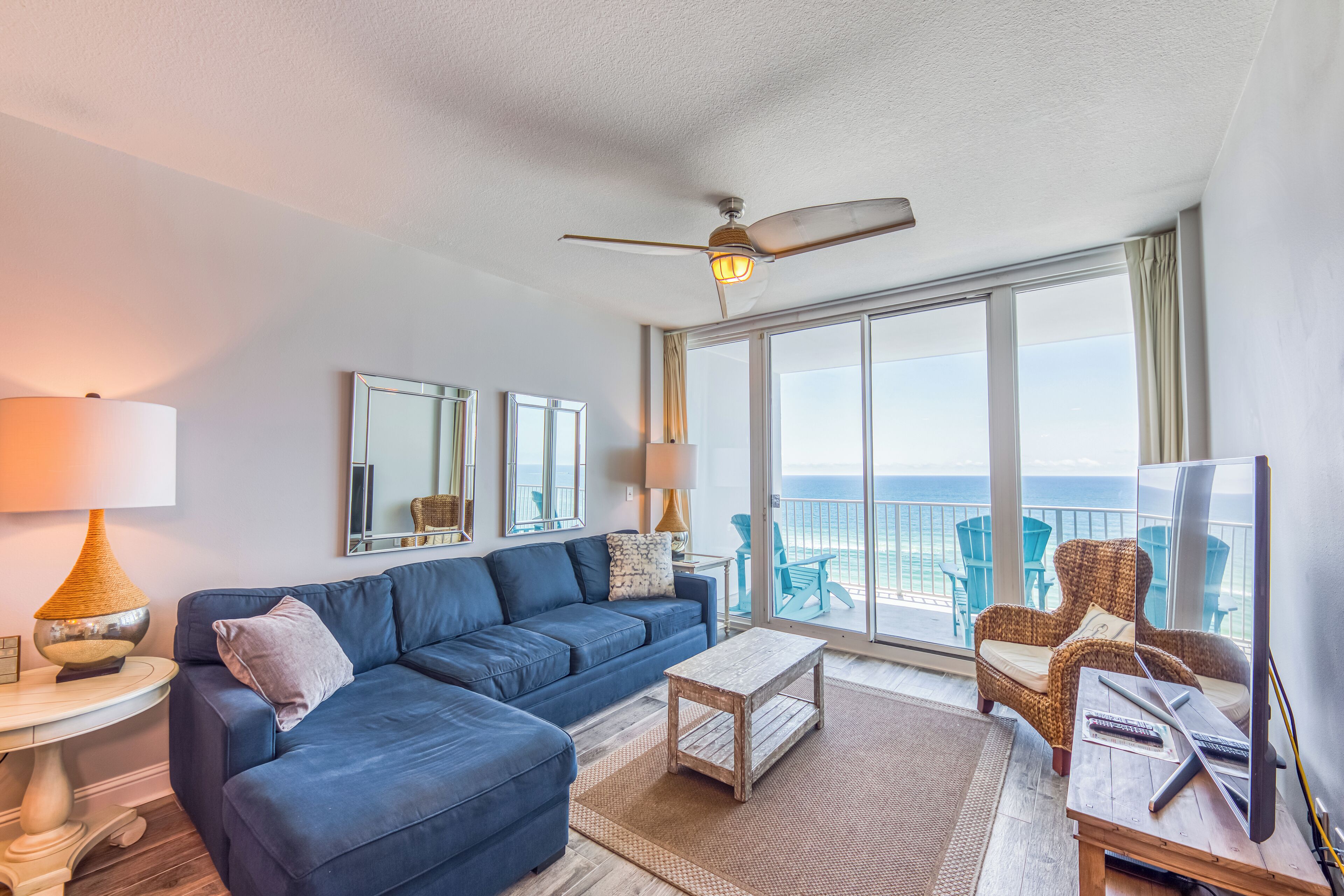 A modern living room with a blue sofa, a wooden coffee table, and a ceiling fan. There are large windows offering a view of the ocean.