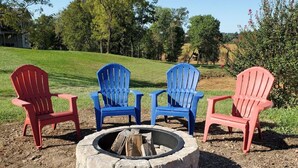 Una chimenea con cuatro sillas Adirondack en rojo y azul, rodeada por un césped con árboles y un edificio al fondo.