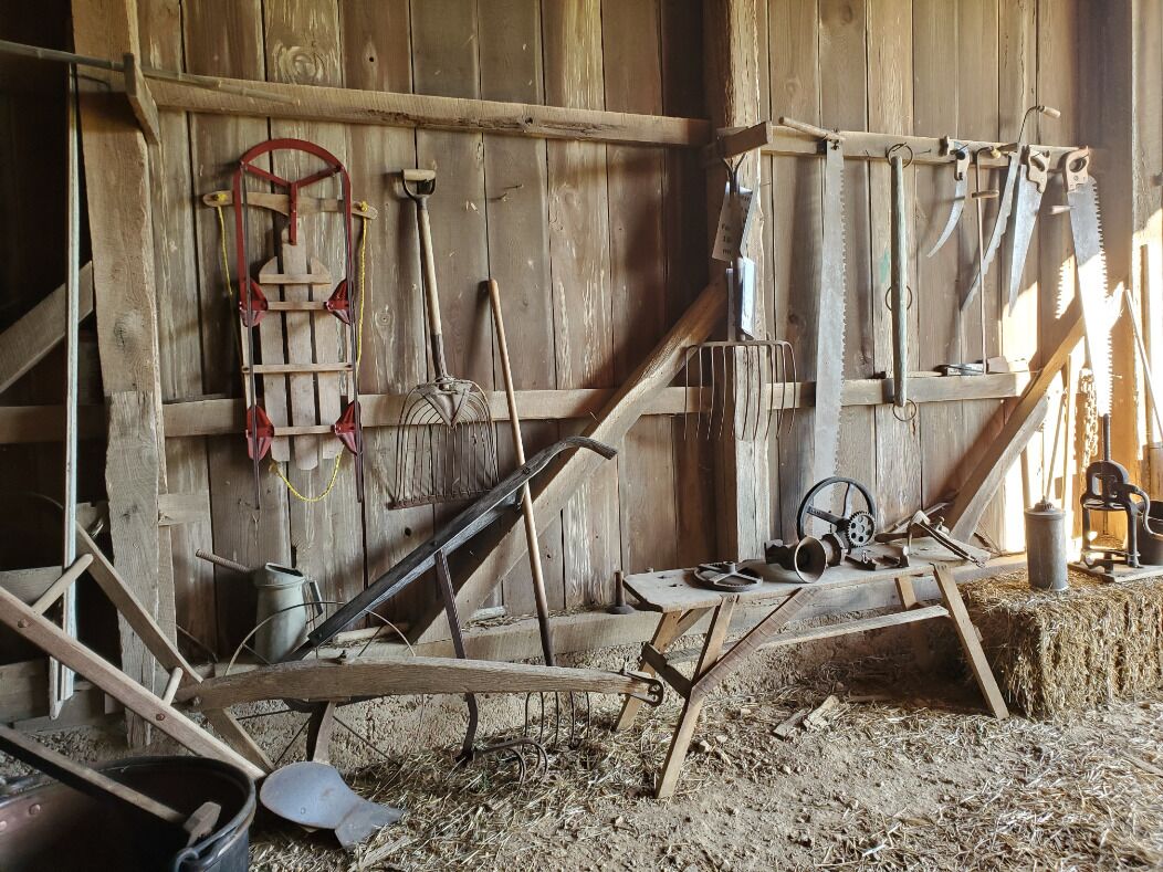 A rustic barn interior with wooden walls, hay on the floor, and various farming tools like a plow, a saw, and a hand pump.