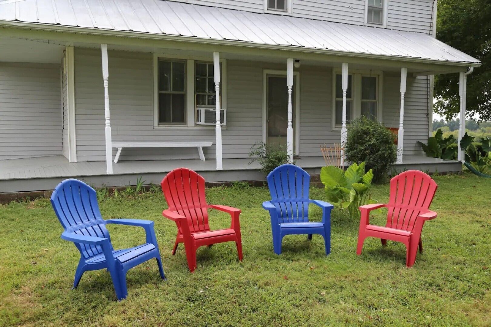 Un porche con cuatro sillas Adirondack en azul y rojo, un banco y una casa con techo de metal.