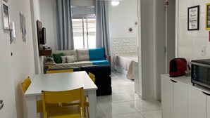A modern living room with a white sofa, a dining table with yellow chairs, a kitchen area with a microwave, and a window with curtains.