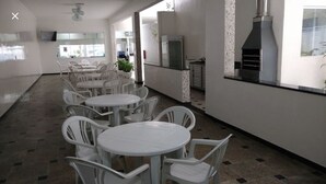 A clean, modern interior with white plastic chairs and tables arranged in a linear fashion, a kitchen area with a built-in stove, and a television mounted on the wall.