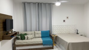 A compact bedroom with a bed, a sofa with a blue throw, a TV on a wooden shelf, and a ceiling fan.
