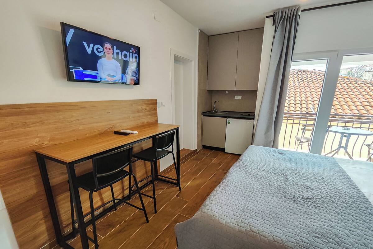 Ein Hotelzimmer mit einem Holztisch, zwei schwarzen Stühlen, einem Bett mit einer Patchworkdecke, einem an der Wand befestigten Fernseher, einer Küchenzeile und einem Balkon mit Blick auf ein Ziegeldach.