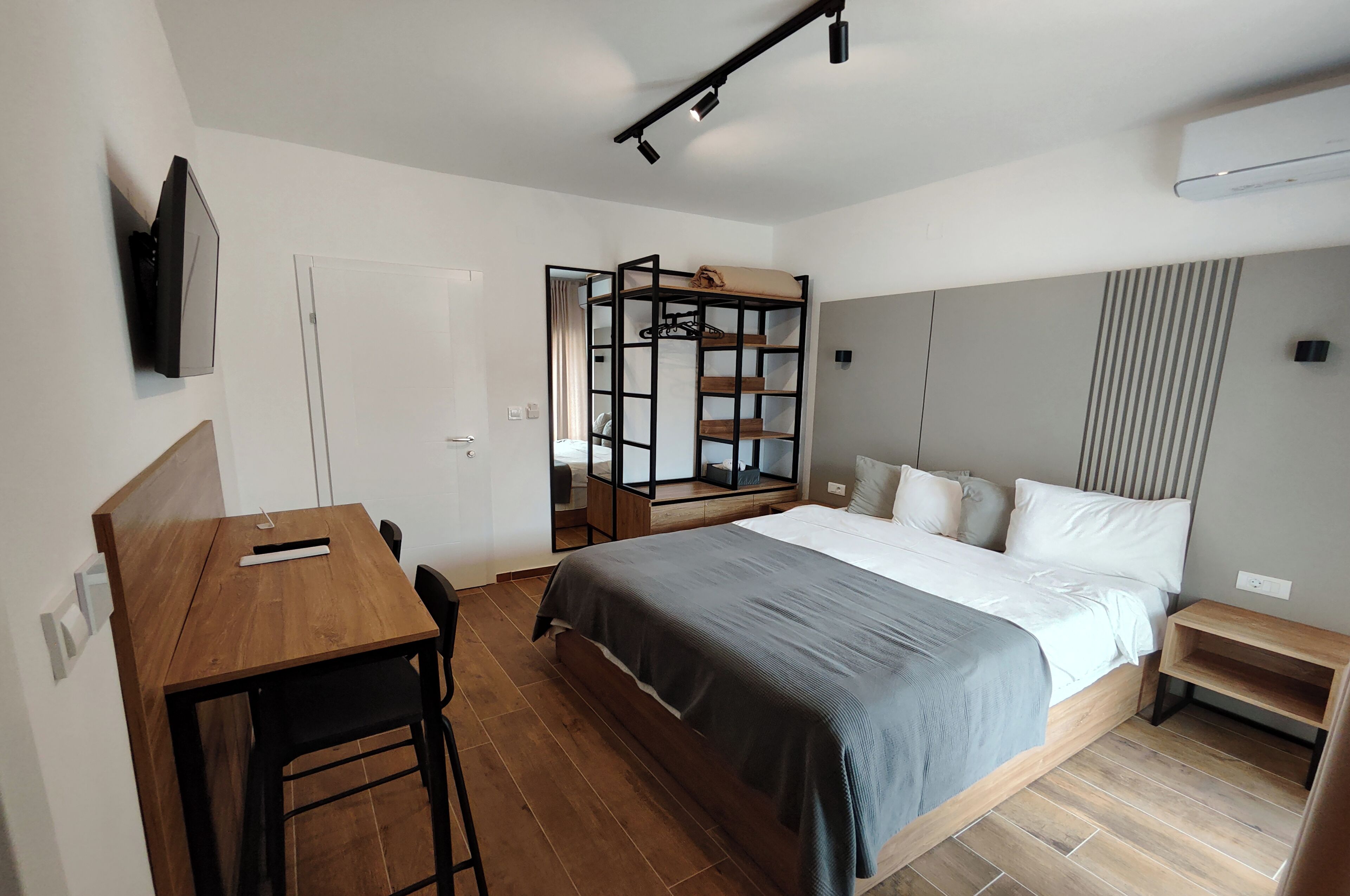 Un dormitorio moderno con un escritorio de madera, una cama con colcha gris, un televisor montado en la pared y un armario empotrado con espejo.