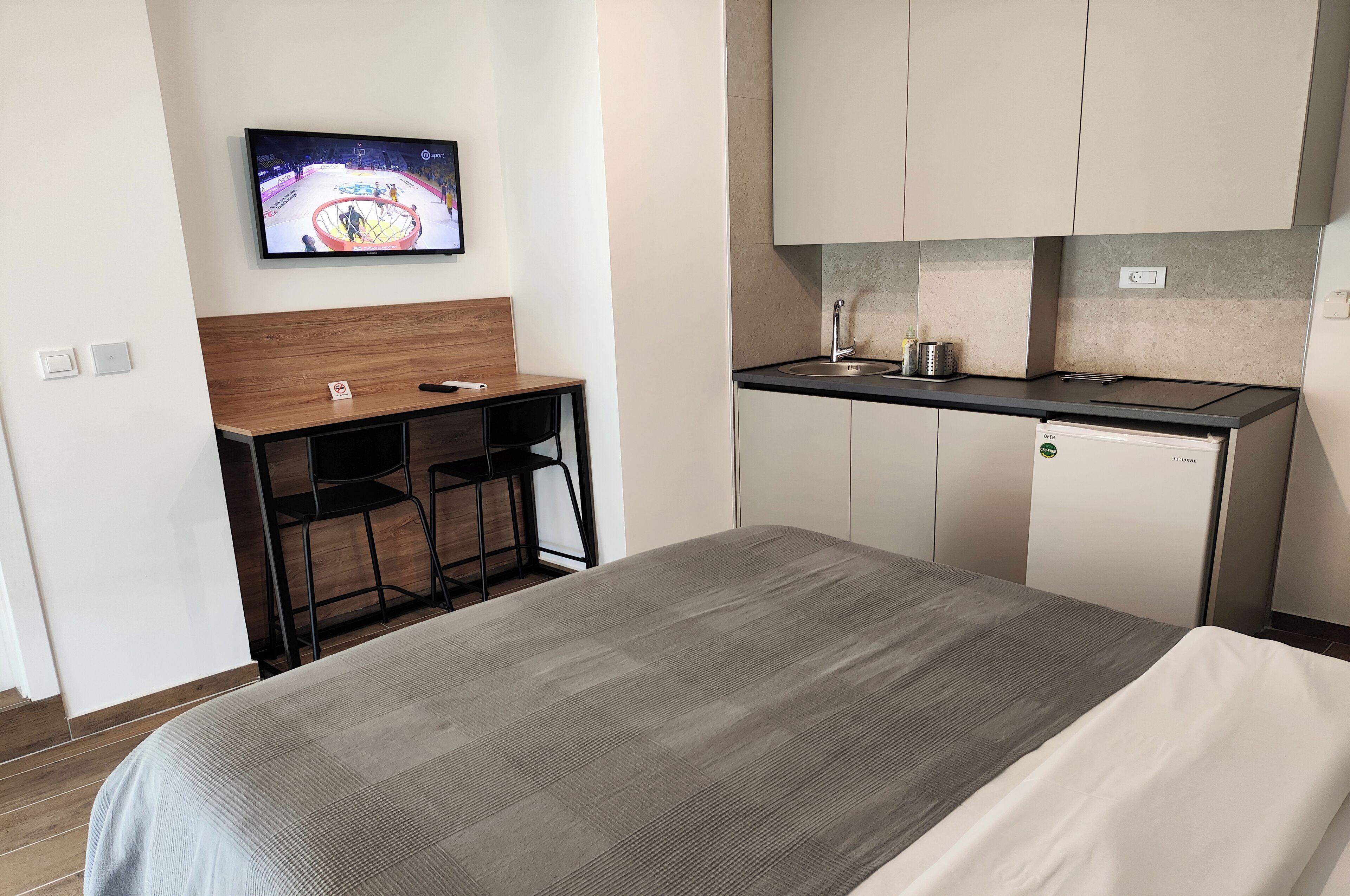 Hotelkamer met een bed, een bureau met stoel, een aan de muur bevestigde televisie, een kitchenette met een koelkast en kastjes.