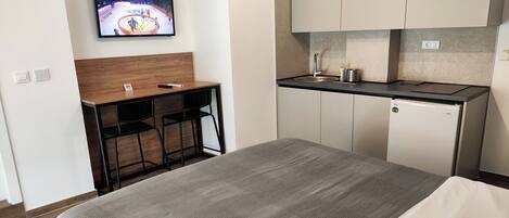 A hotel room with a bed, a desk with a chair, a TV mounted on the wall, a kitchenette with a fridge, and cabinets.