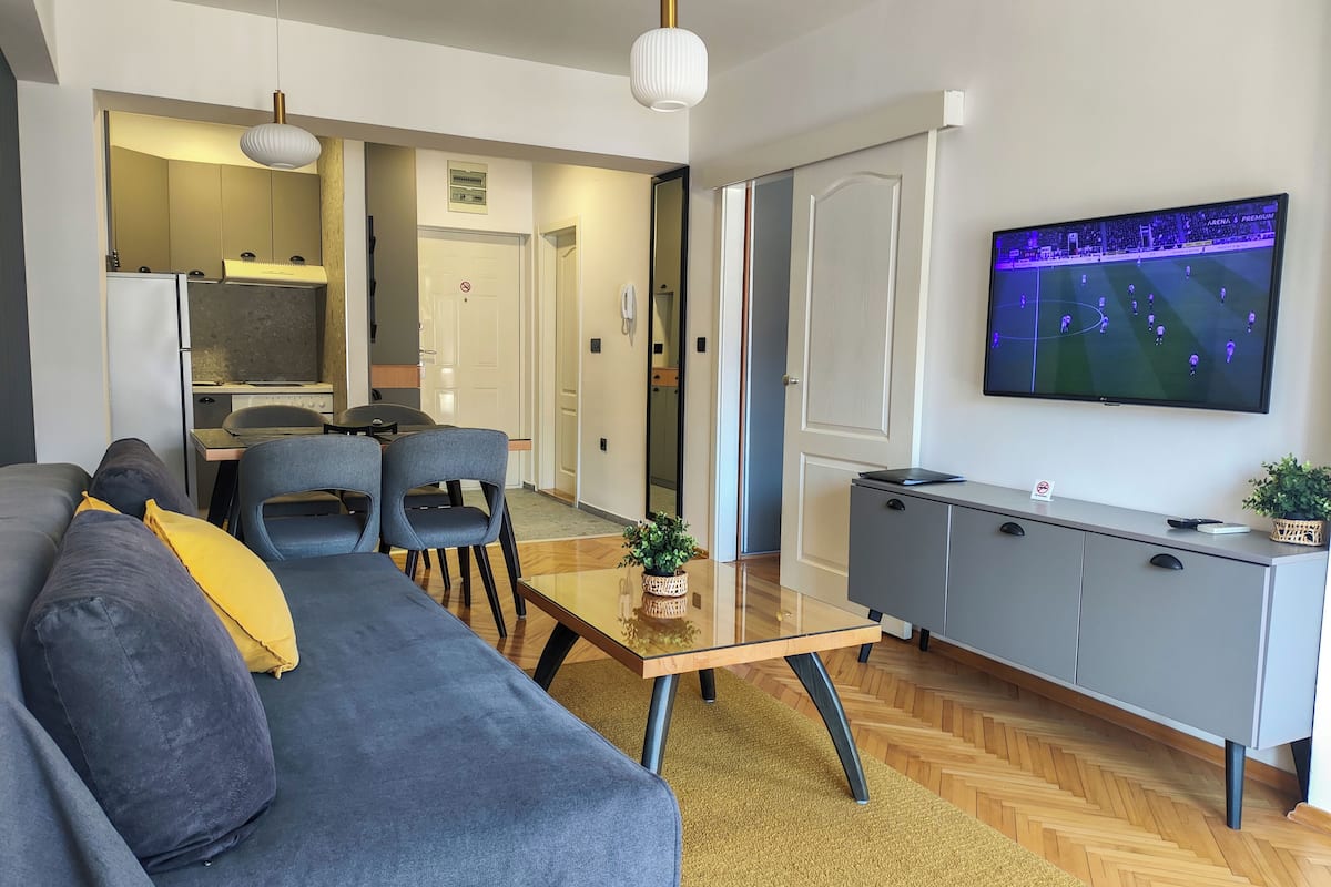 Una sala de estar moderna con sofá, sillas, una mesa de centro, televisor, zona de cocina y mesa de comedor.