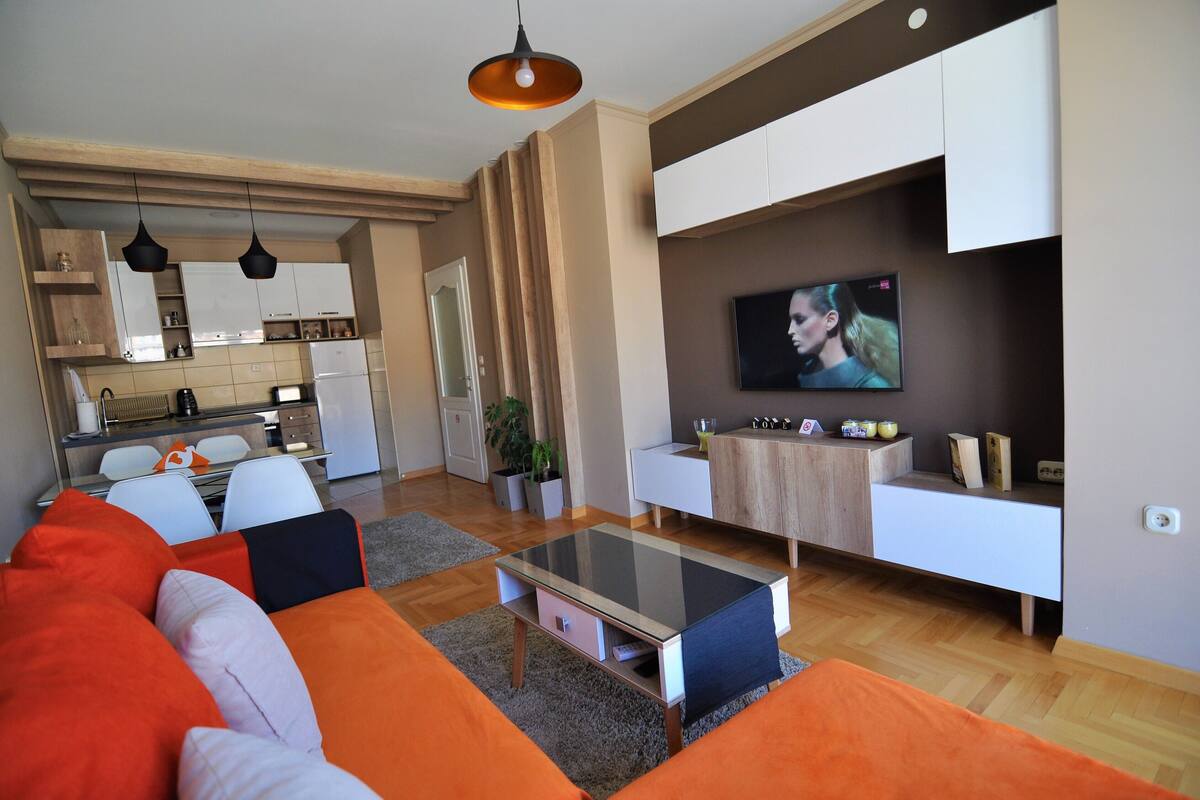 Una sala de estar moderna con una mesa de centro de vidrio, un sofá naranja y blanco, y un televisor montado en la pared.