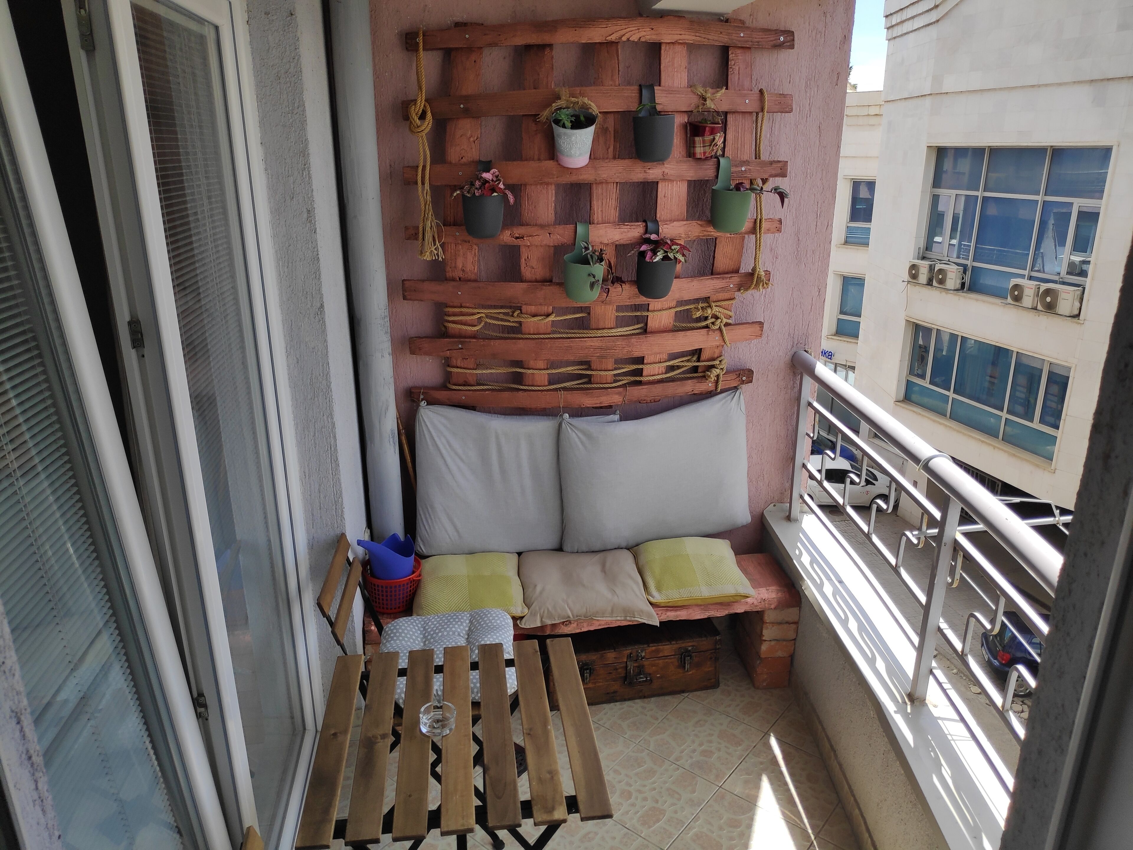 A balcony with a wooden bench, potted plants, and a cushioned seating area.