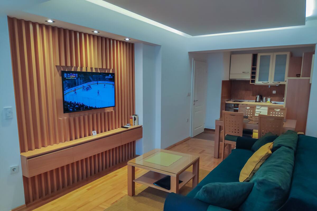 Una sala moderna con televisor de pantalla plana montado en una pared de paneles de madera, un sofá verde, una pequeña mesa de centro de madera, y una zona de cocina con armarios de madera y una mesa de comedor.