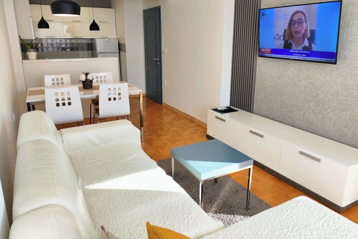 Una sala de estar moderna con un sofá blanco, un televisor montado en la pared y un comedor con mesa y sillas.
