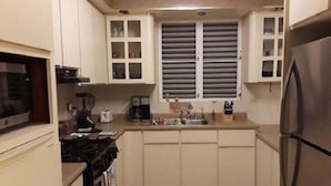 Cocina compacta con estufa, pileta y refrigerador. Hay alacenas sobre la encimera y un ventanal con persianas.