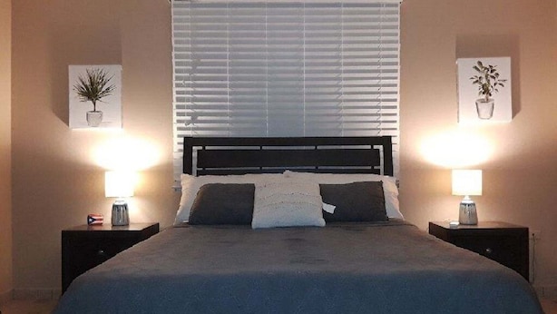 Una cama bien hecha con un cabecero oscuro, flanqueada por dos mesitas de noche con lámparas, en una habitación con paredes color beige y un ventanal con persianas.